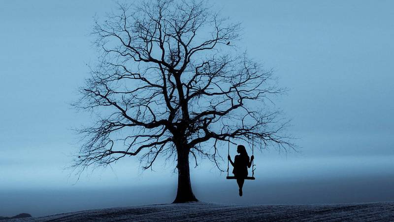 Siluett av kvinna som gungar från ett träd där löven fallit av, mot en molntung blåtonad himmel.