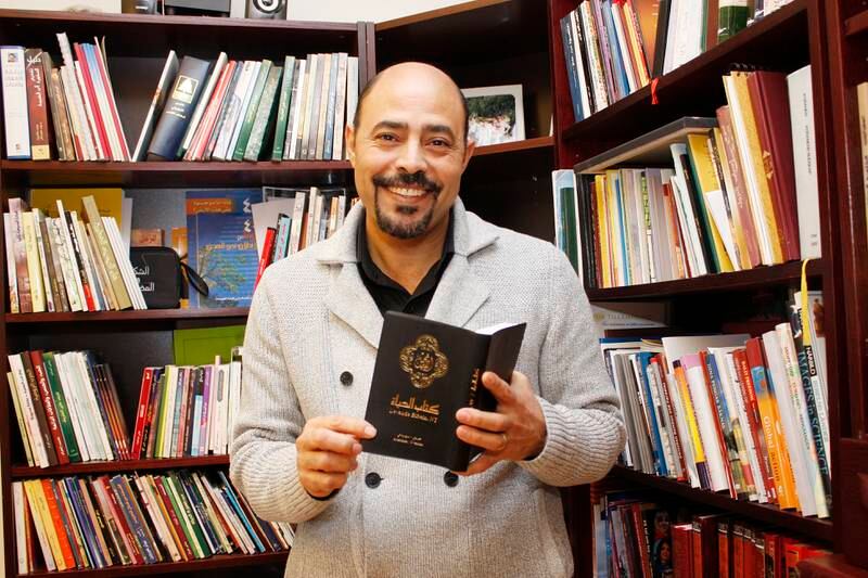 Merzek Botros håller en bibel på arabiska, står bland fulla bokhyllor.