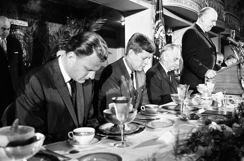President John F. Kennedy böjer sitt huvud under bönen vid bönefrukosten 1961. Till vänster i bild syns evangelisten Billy Graham.
