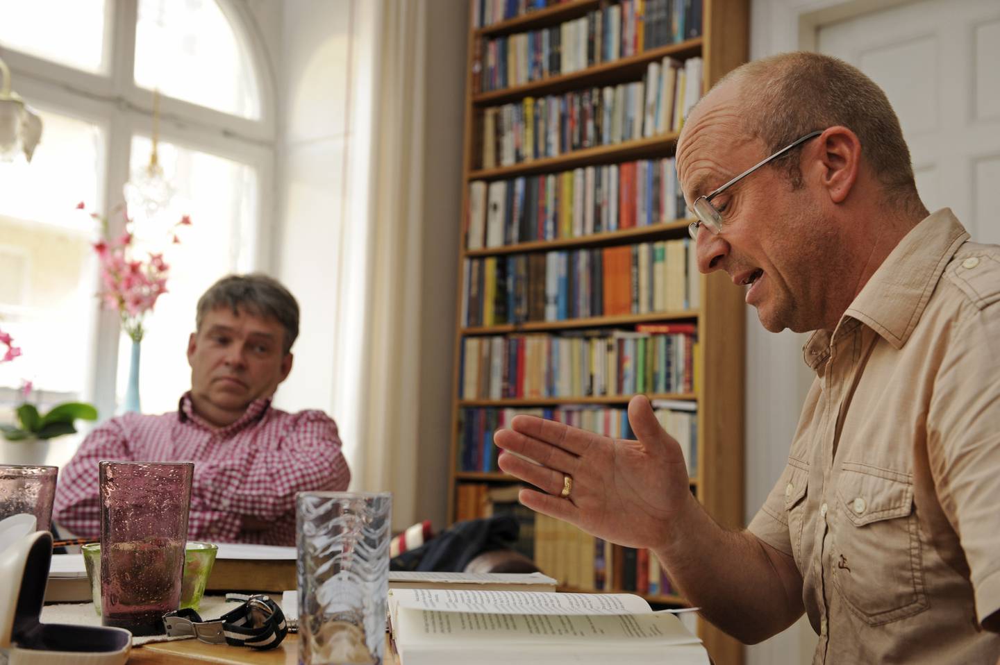 Stefan Swärd och Jonas Gardell sitter vid ett matbord och äter lunch och diskuterar tro i ett rum med bokhyllor.