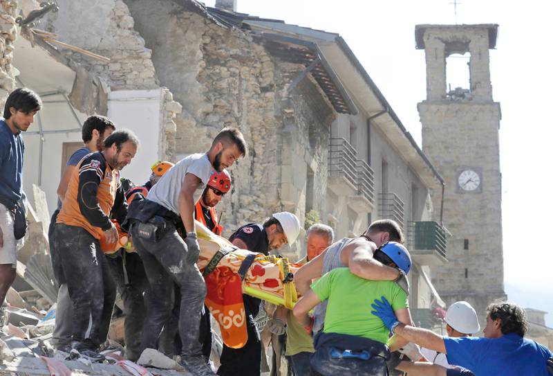 Ett offer bärs på en bår från en kollapsad byggnad efter den svåra jordbävningen. I bakgrunden syns stadens skadade 1400-talskyrka.