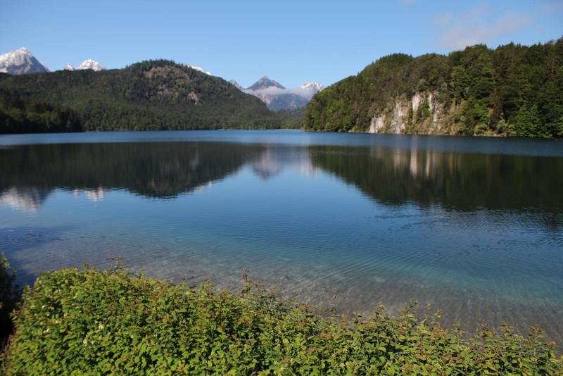Vykortsvy. Spegelblankt vatten, dramatisk natur och höga berg. Många av alptopparna som bildar fond ligger i grannlandet Österrike.